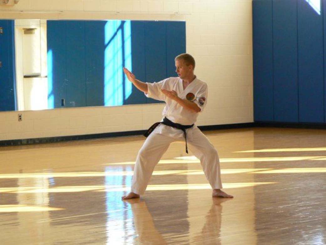 Man wearing a gi and black belt stands in a wide-leg position facing the left with arms extended, practicing martial arts in a gym.