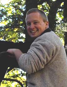 Man with light skin and very short brown hair stands smiling in front of trees. He is wearing a tan sweater.