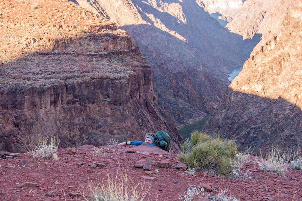 Red rock ledges take up most of the image, and a man wearing a blue shirt, blue bandana, carrying a large green backpack, peers over the ledge as he climbs down the mountainside.