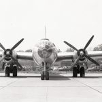 Front view of large World War II aircraft is parked behind hangar.