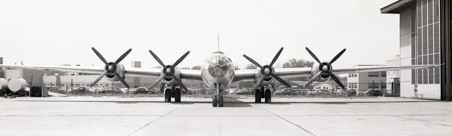 Front view of large World War II aircraft is parked behind hangar.