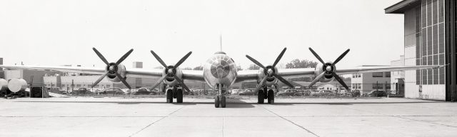Front view of large World War II aircraft is parked behind hangar.