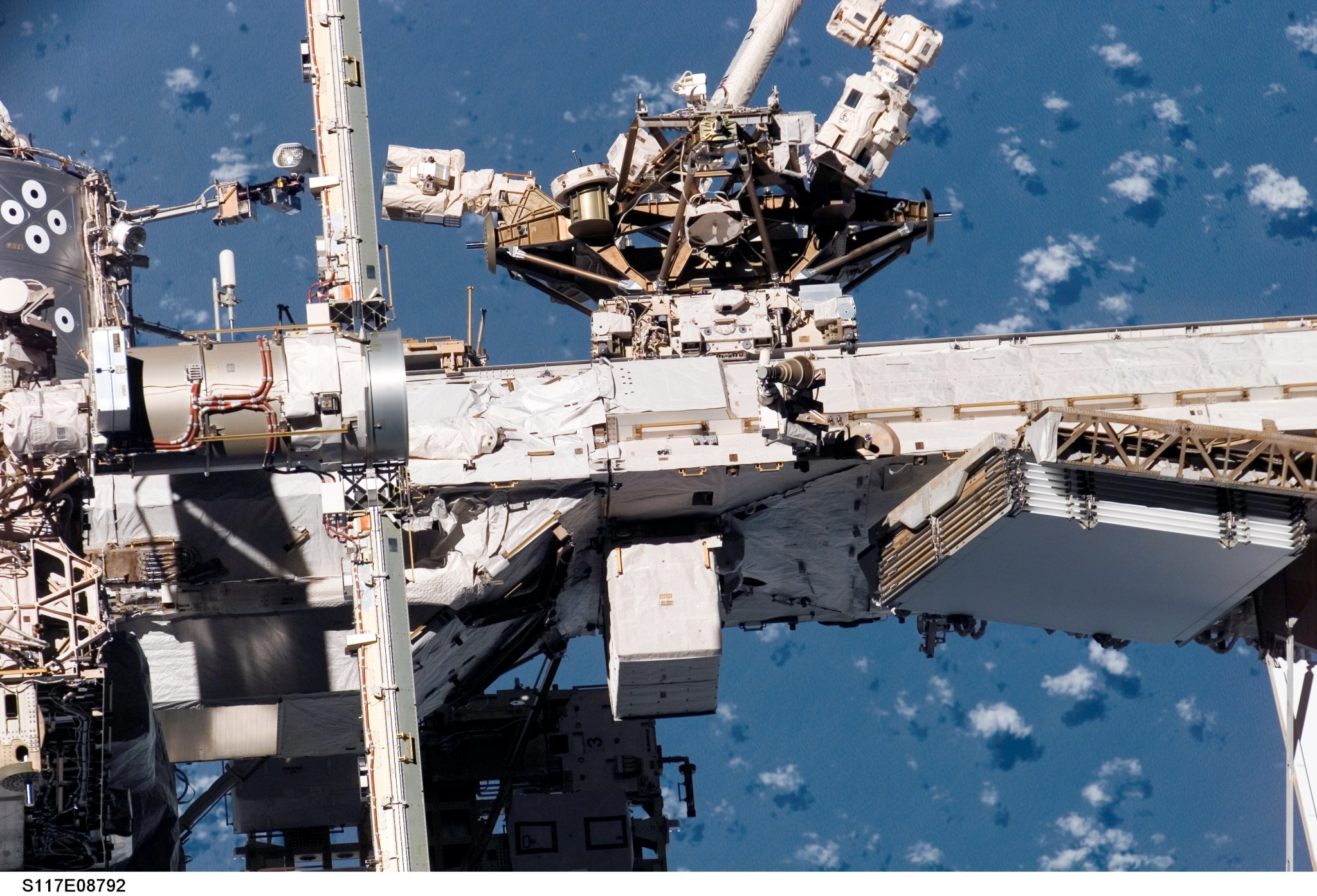 Exterior view of portion of ISS, with several white and silver technical components, with blue-hued Earth in background