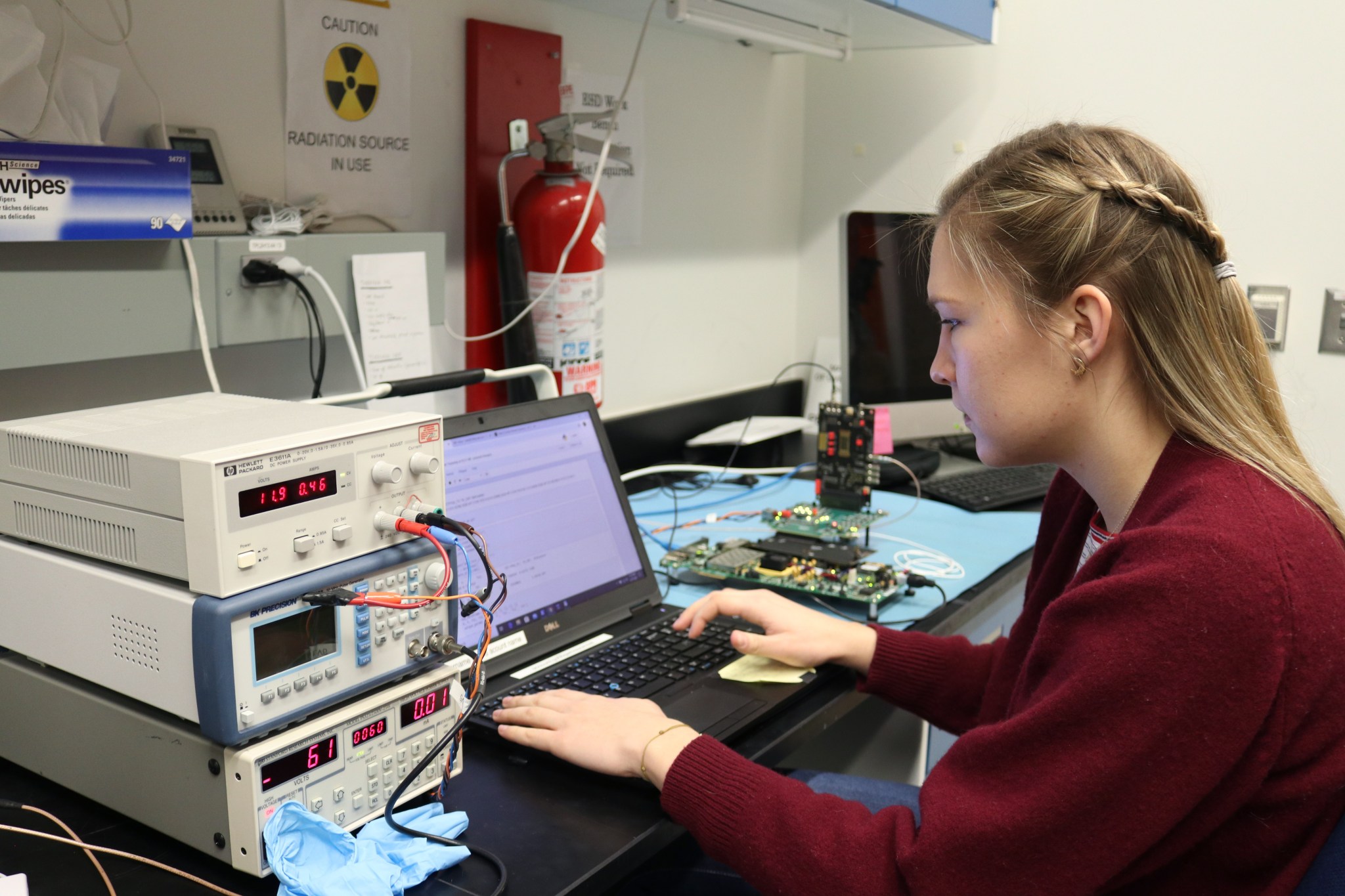 Woman with fair skin and light brown hair works on a laptop at a desk with machines on it. She looks focused. She is wearing a red sweater.