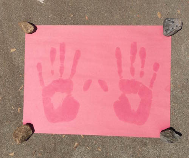A faded sheet of pink construction paper with two red handprints. A small rock holds down each corner of the paper.