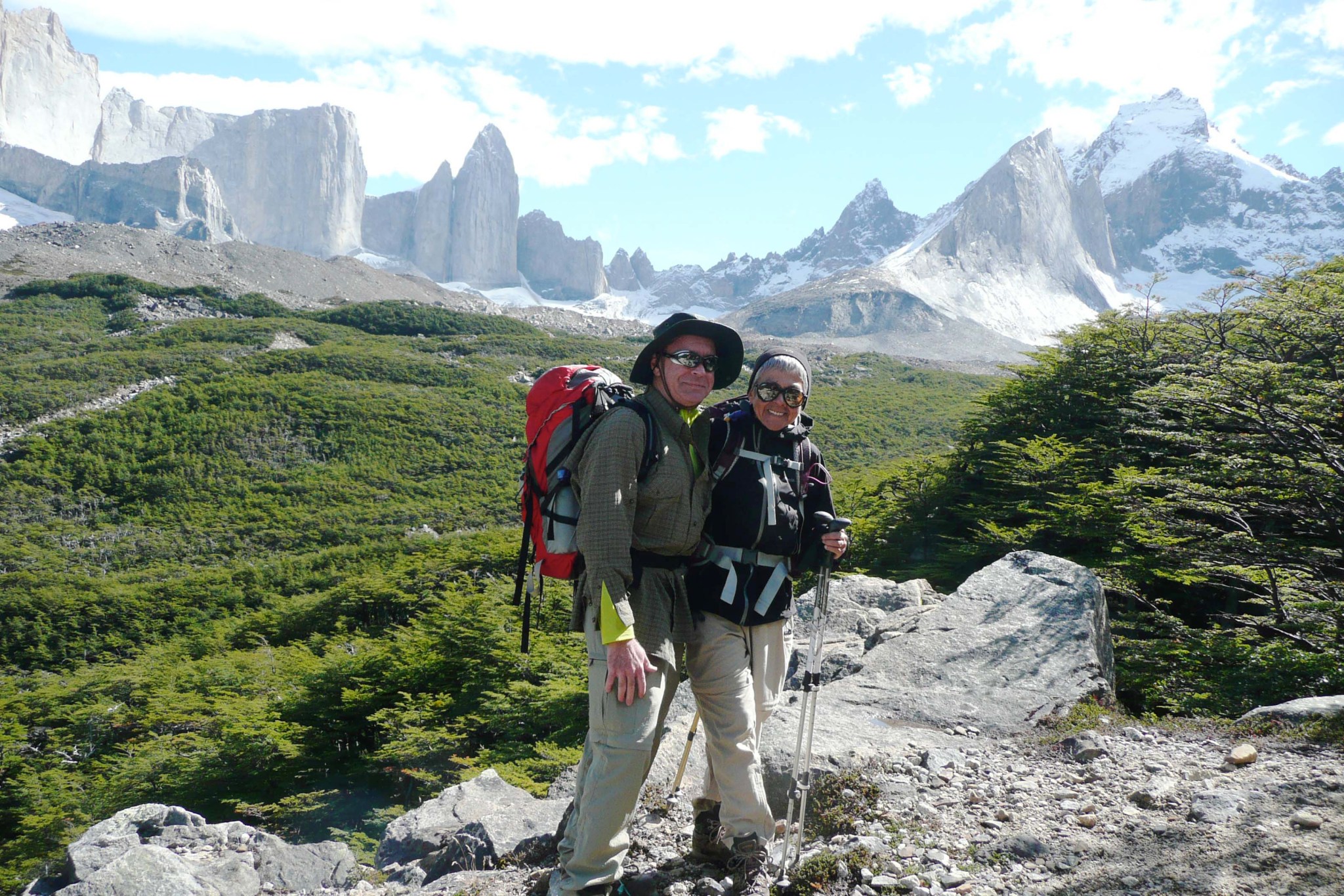 Man and woman in hiking clothes, hats, and backpacks, hold hiking poles and pose for a photo on a trail with mountains and greenery behind them. 
