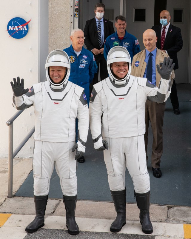Two astronauts wearing white flight suits and helmets while waving to the crowd.