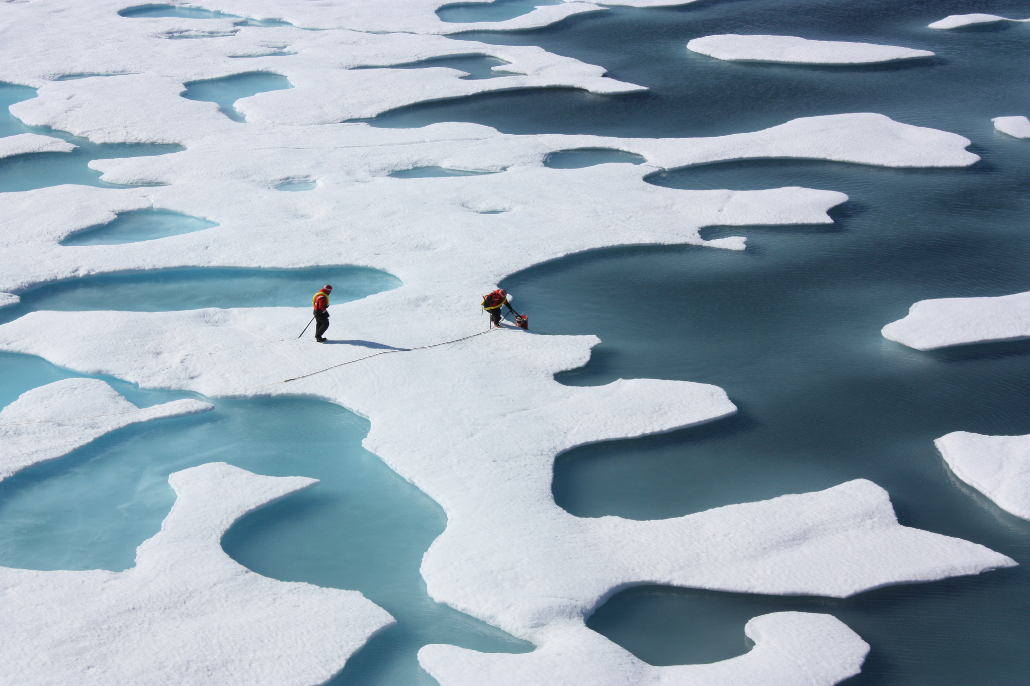two people stand on ice sheets, some parts are melted. Blue water is visible everywhere the ice is not