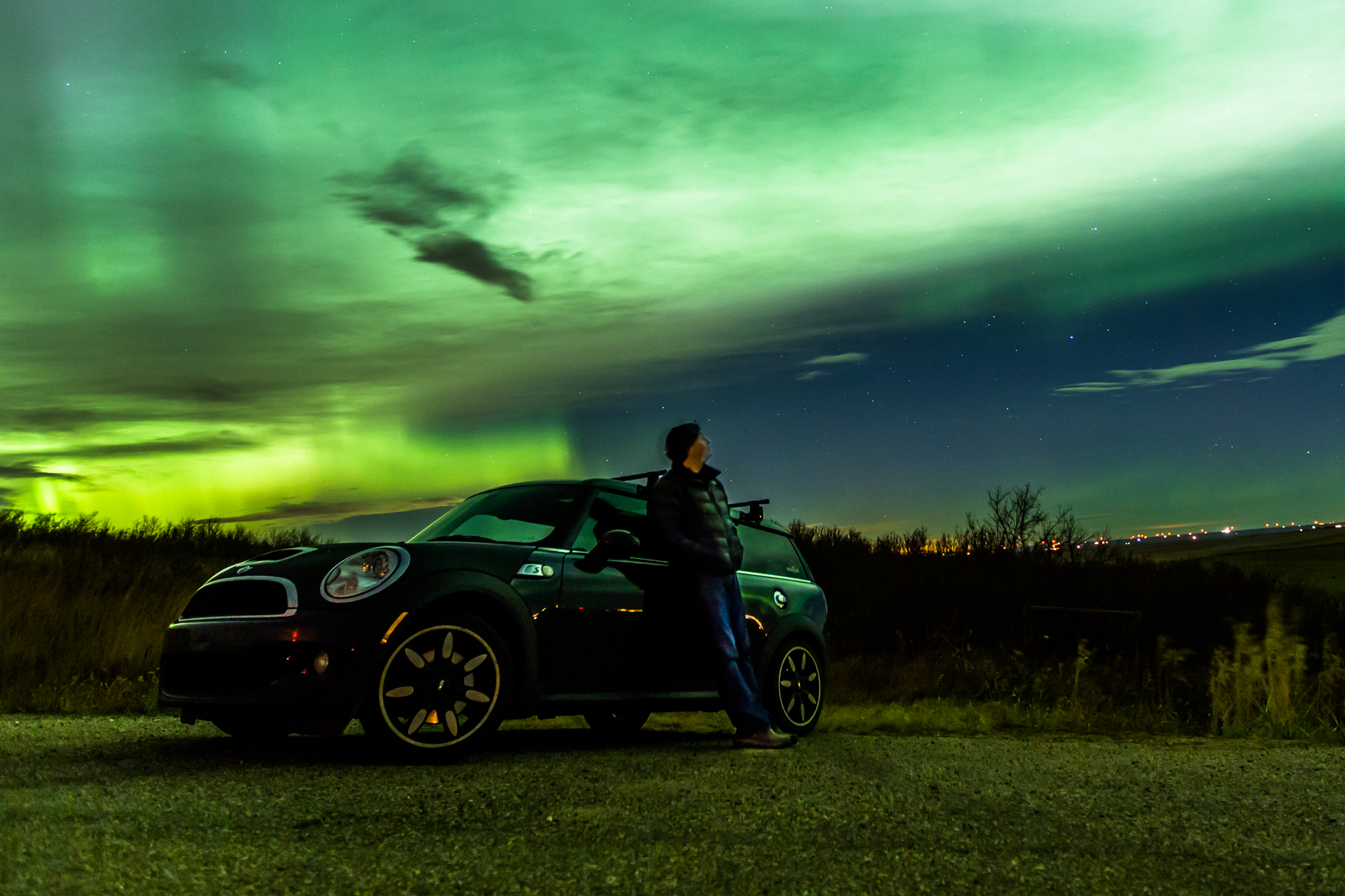 Aurora chaser Chris Ratzlaff leans against a car while looking up at an aurora-filled night sky