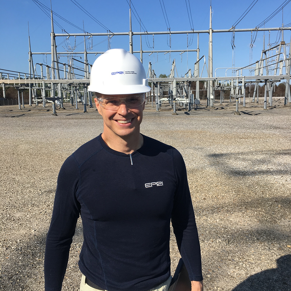 Bob Arritt smiles at the camera and stands in front of electric power equipment while wearing a white hard hat and safety goggles.