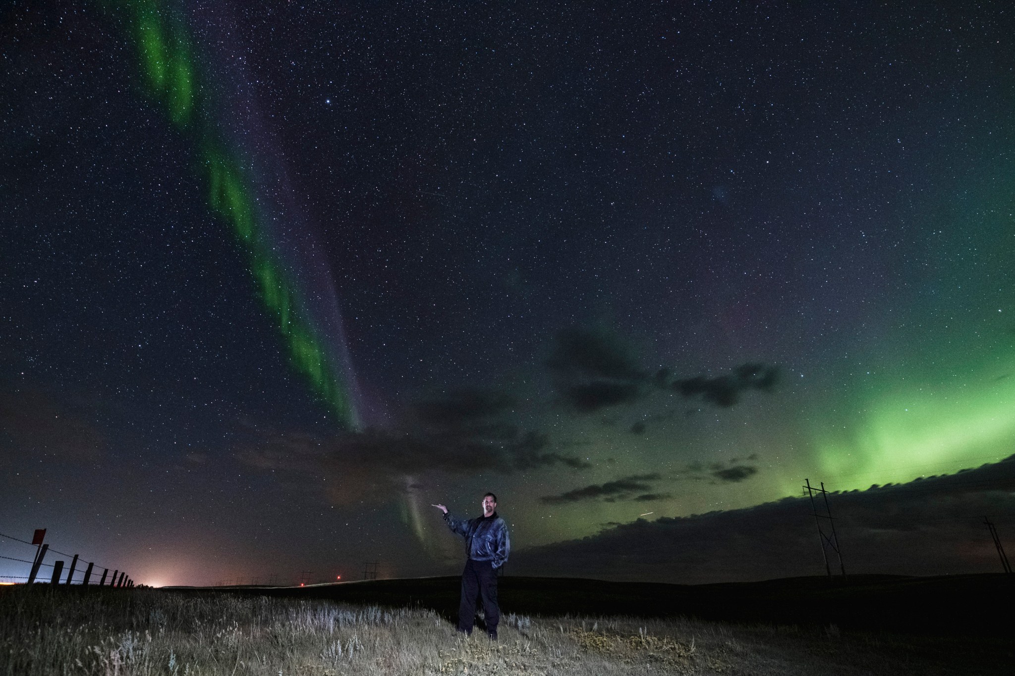 Notanee Bourassa stands in a field under the night sky with a purple and green aurora structure on the left and a green glow in the distance on the right.