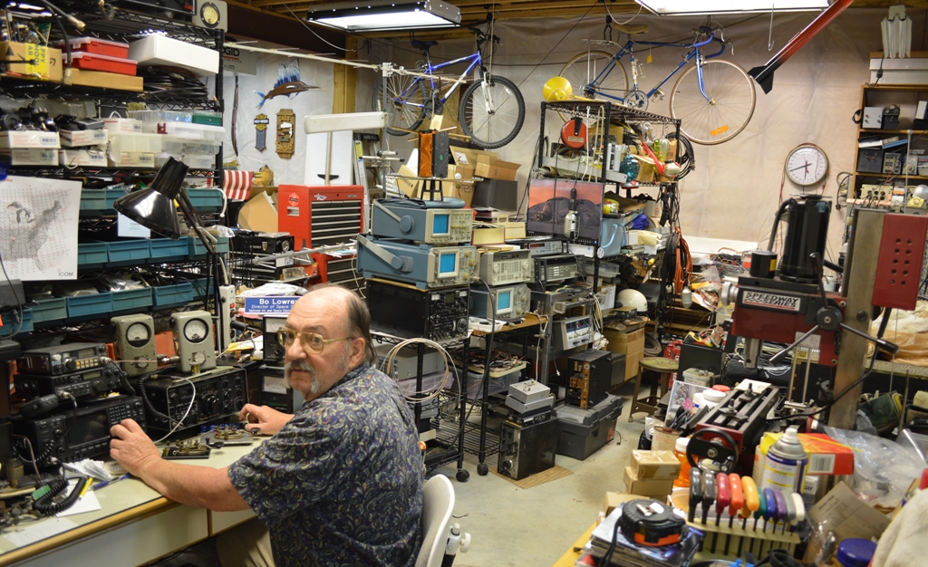 Bo Lowrey sits in front of some radio equipment, with his left hand on a knob, while turning his head to look at the camera over his left shoulder.