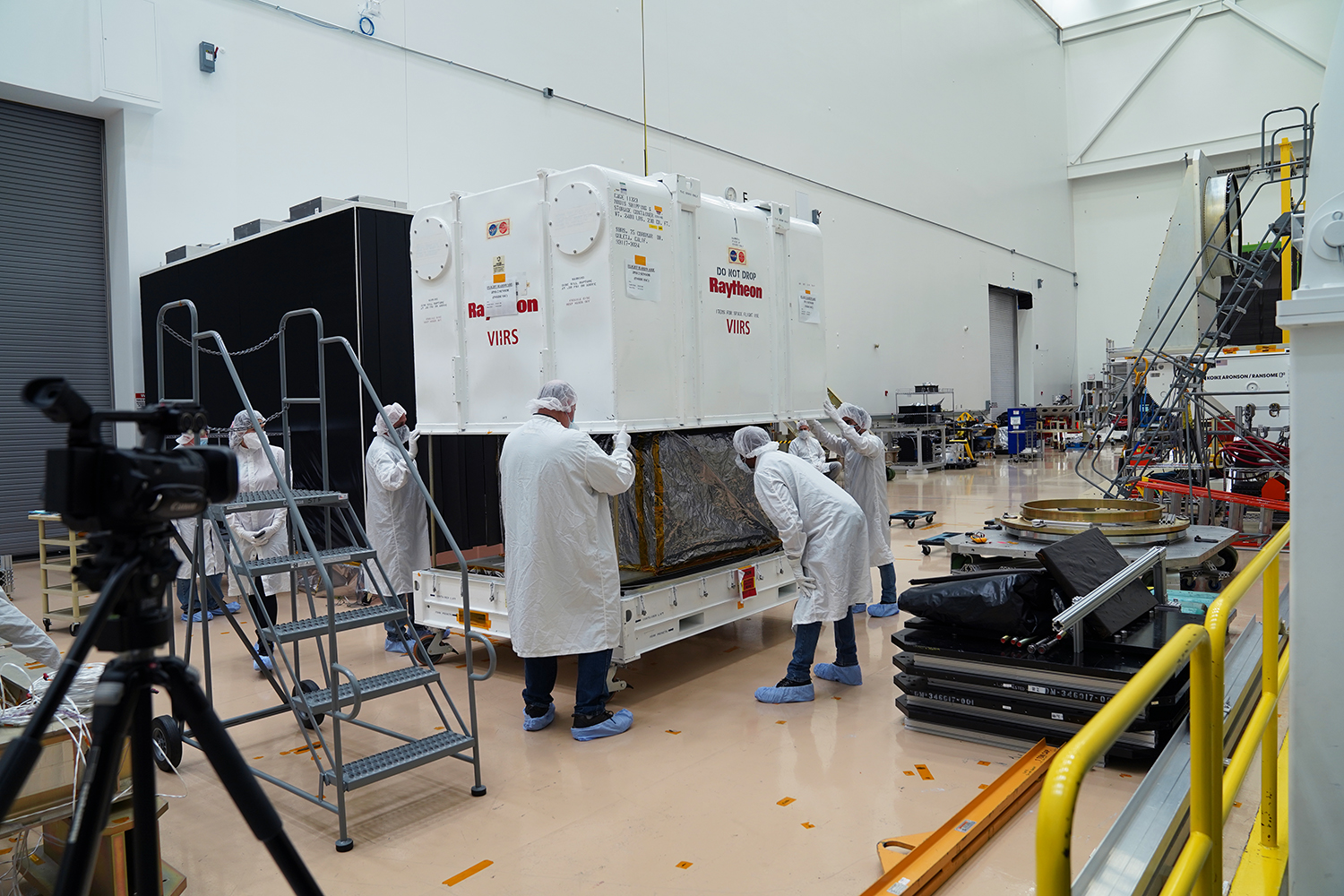 Engineers in white lab coats and hair nets help remove a large white box from a rack carrying the VIIRS instrument for JPSS-2. The instrument is wrapped in silver foil.
