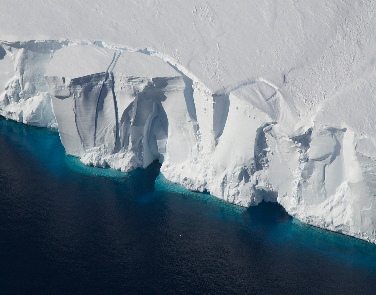 Photo of an Antarctic ice shelf. A tall, mostly sheer cliff of white ice rises out of a dark blue ocean.