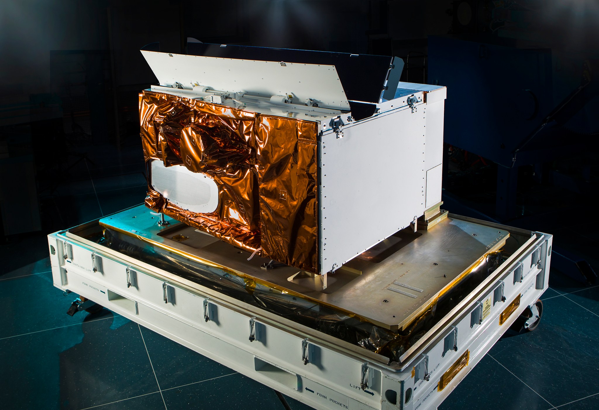 The VIIRS instrument in a dark room. It is lit so its bronze-foil wrapped side glistens. The other side are mostly a dull white color. It's roughly rectangular.