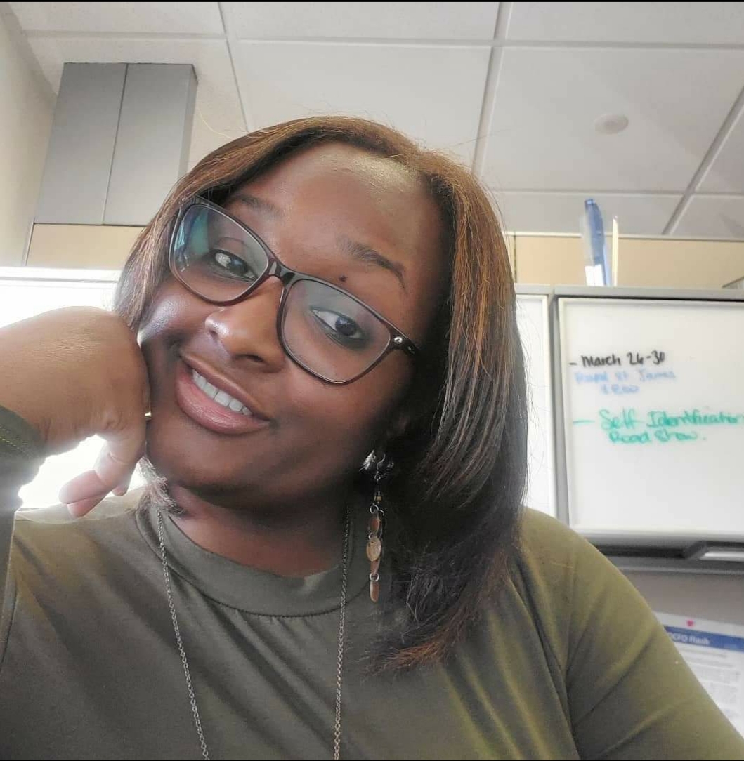 a Black woman with shoulder length, brown hair, wearing a green shirt and glasses rests her head on her hand and smiles