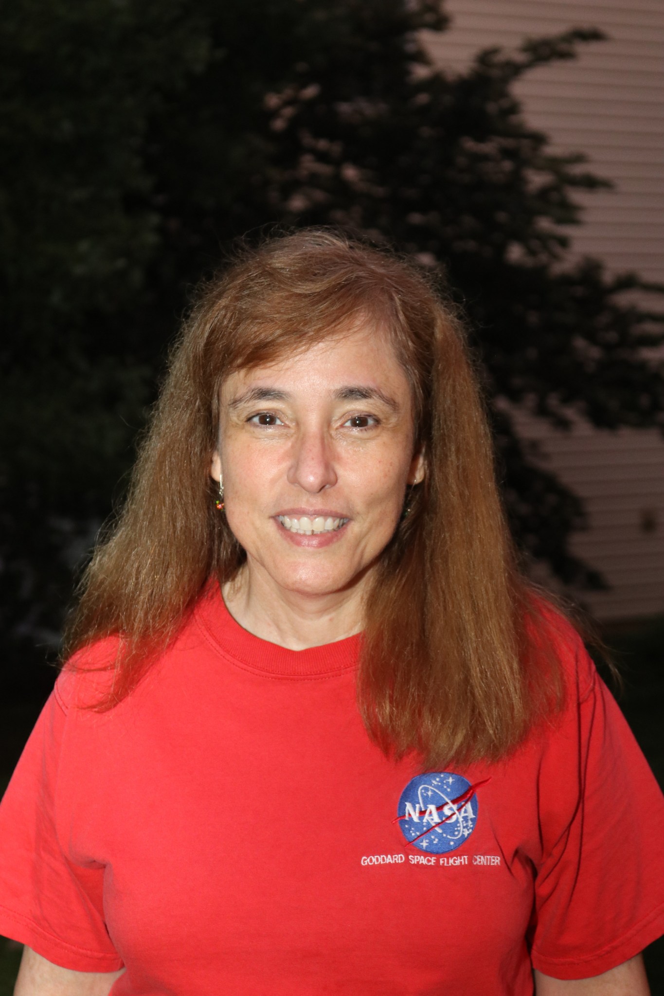 Woman with reddish brown shoulder length hair smiles wearing a red shirt with a NASA logo that says "Goddard Space Flight Center" 
