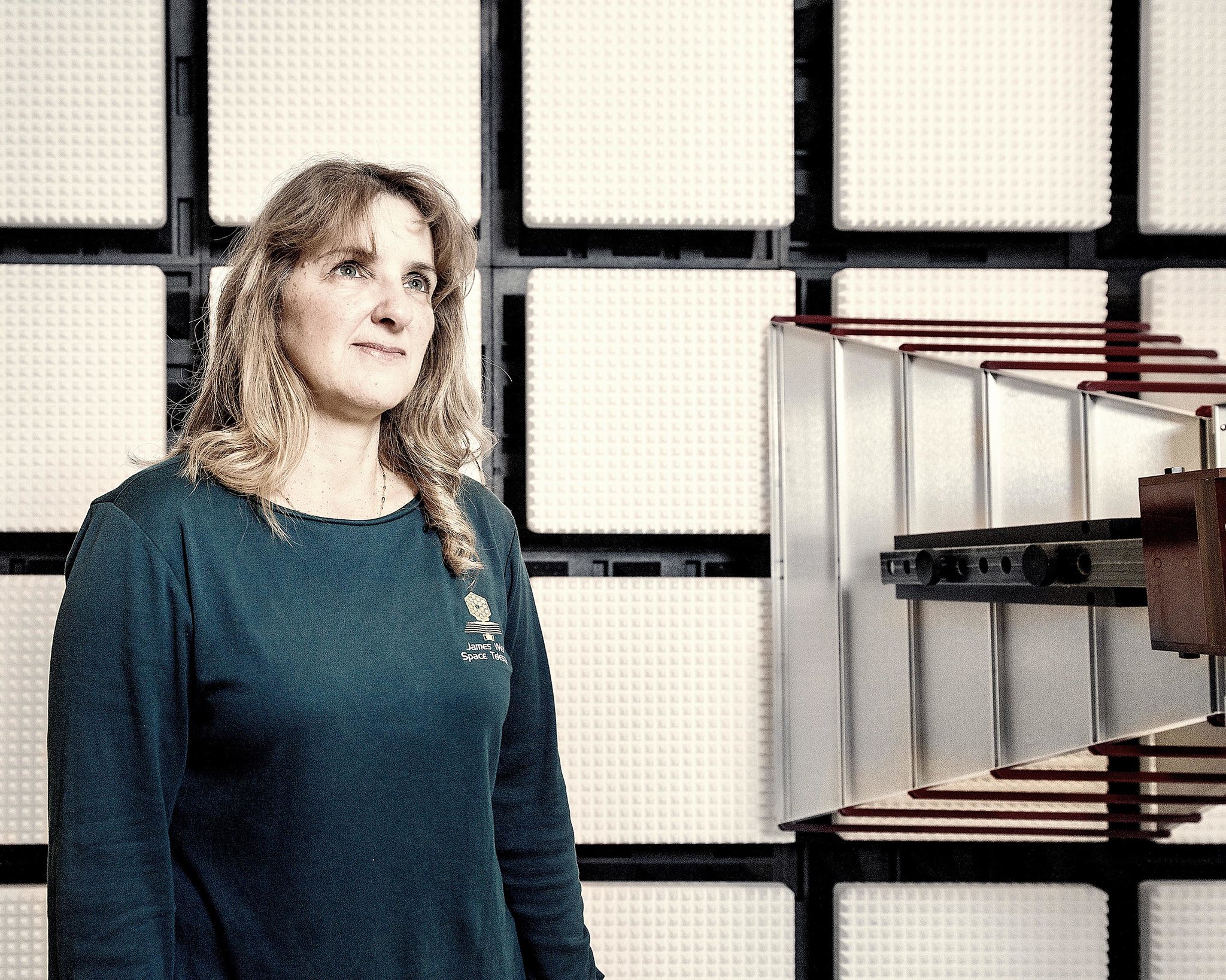 A woman with brown shoulder length hair wears a teal shirt with the Webb telescope logo on the left looks off camera in a portait. The background is a grid of 4x4 blocks.