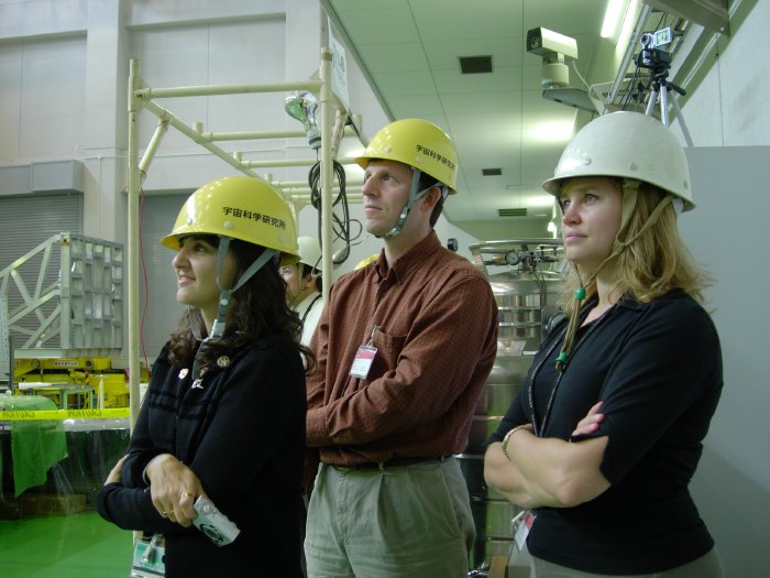 Three people wear hard hats and look off screen to the left. The woman in front, furthest to the left,  has dark, curly hair and wears a black long sleeve shirt and holds a small camera, the man in the middle wears a burnt orange dress shirt with tan pants, and the woman furthest to the right wears a black 3/4s sleeve shirt and has blonde hair. They are standing in a lab. 