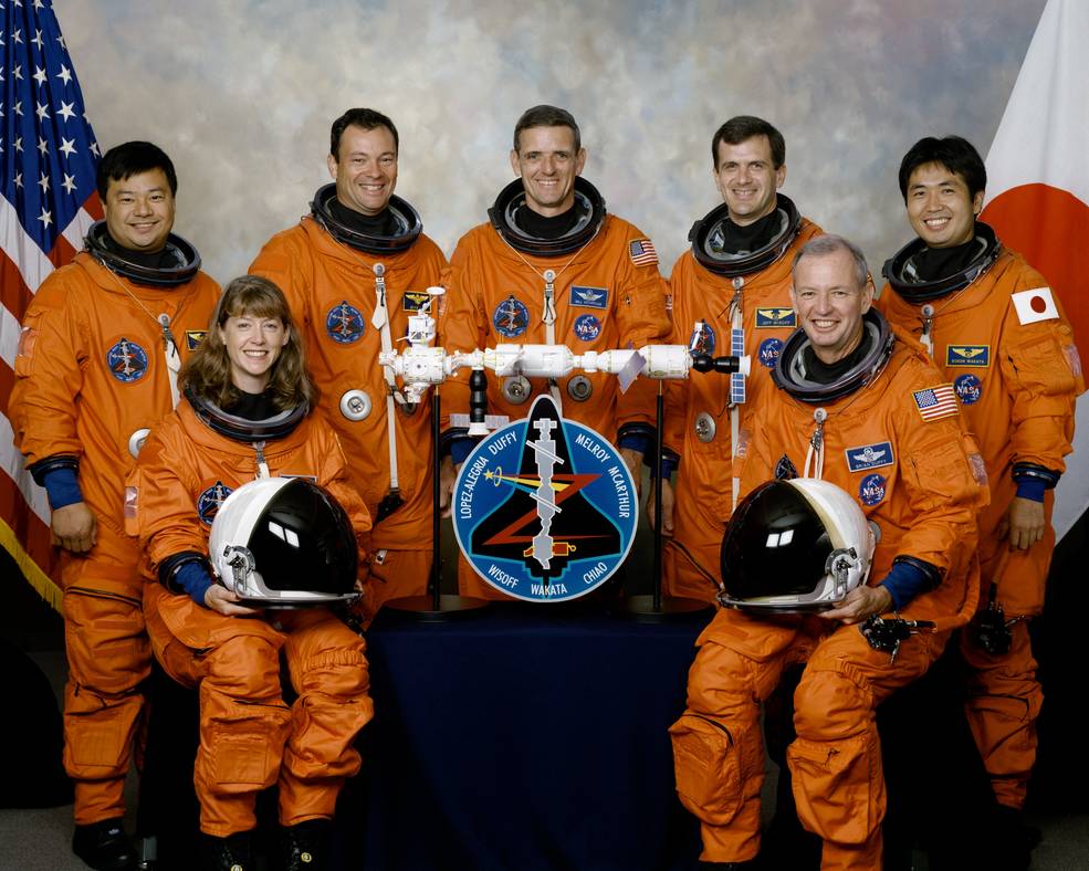 Seven astronauts in orange spacesuits pose in front of US and Japanese flag