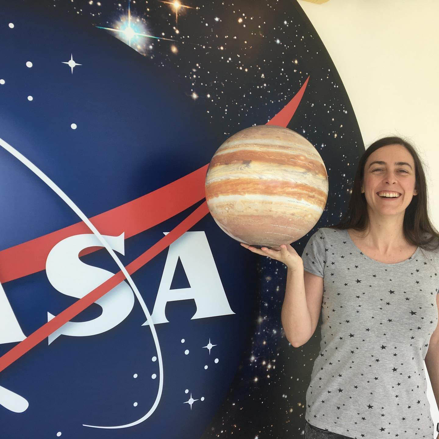 Woman with brown hair, wearing a grey shirt with black stars on it holds a model of planet Jupiter in front of the NASA meatball logo.