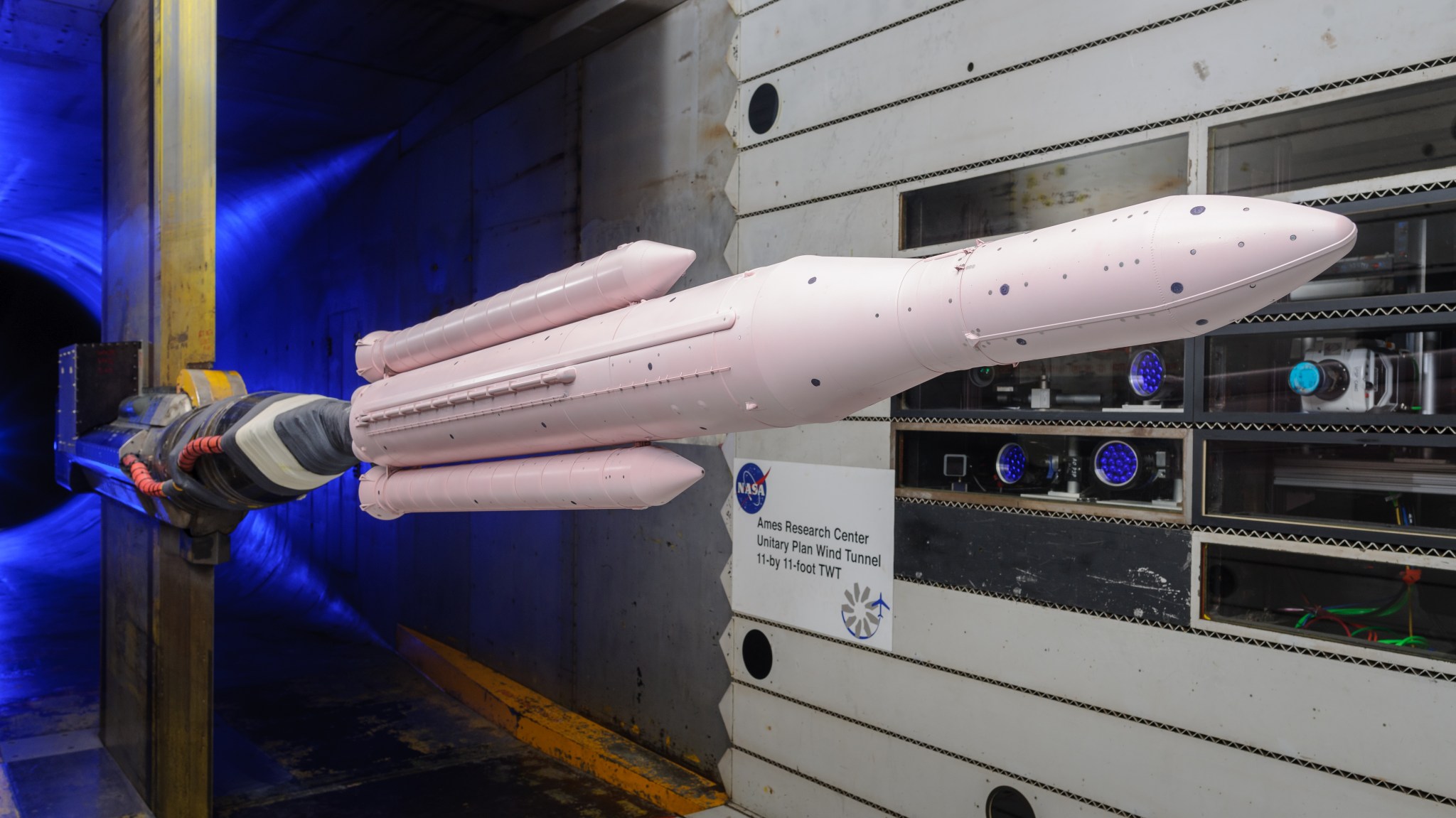 A rocket model painted pink is mounted in a wind tunnel with blue light behind