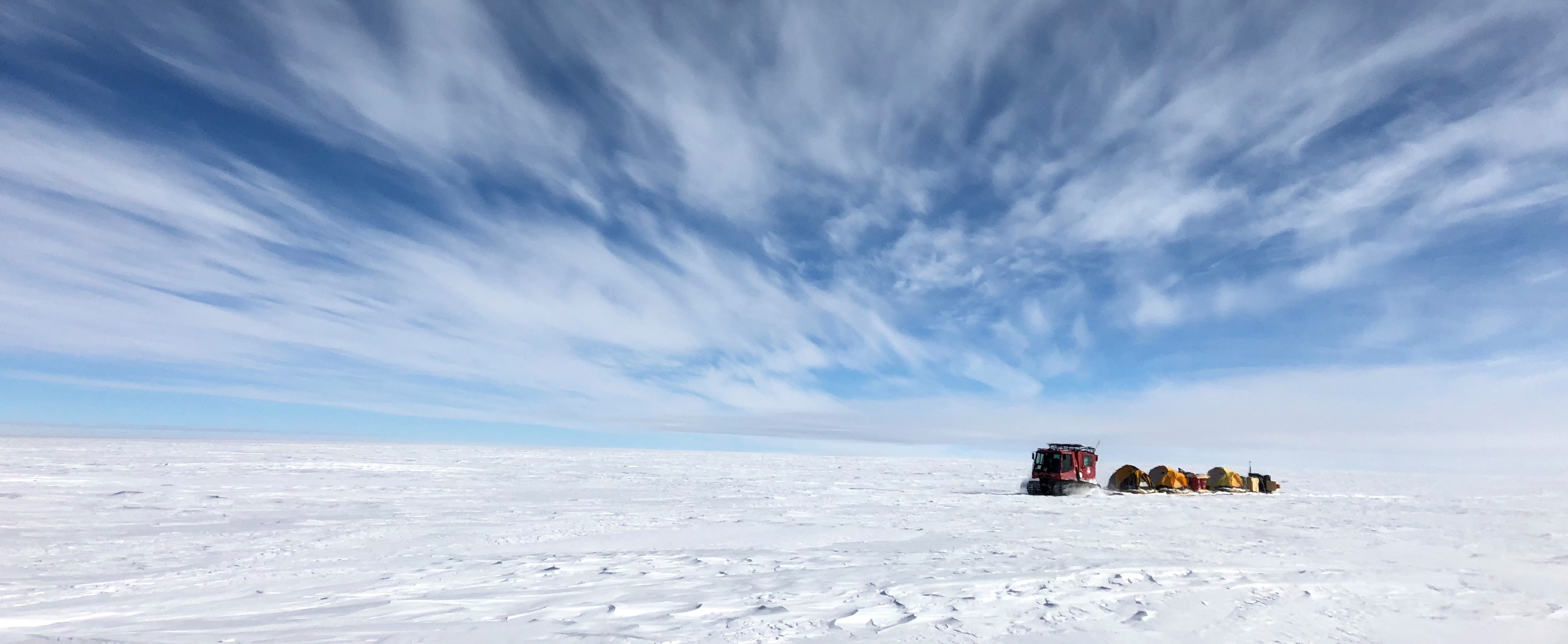 A panoramic photo of the Antarctic landscape, with a large red vehicle towing a chain of supply barges across the windswept white snow. The sky is deep blue with streaky clouds radiating across it.