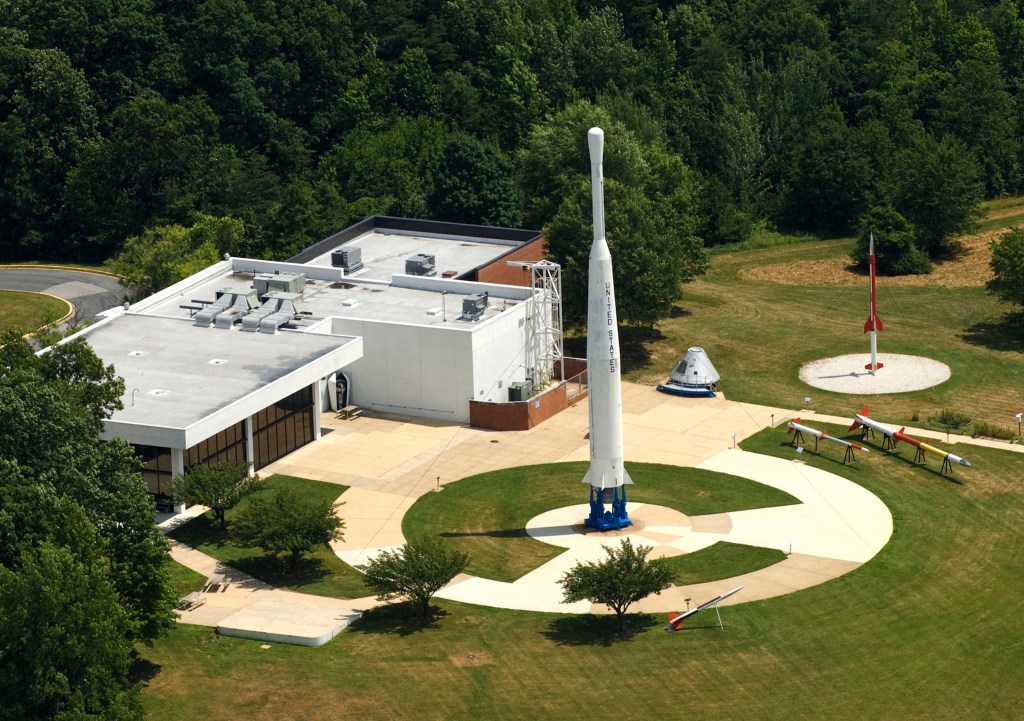 aerial view of a low-slung office building with a circular concrete sidewalk behind it, featuring a tall Delta A rocket in its center, with green grass and trees surrounding
