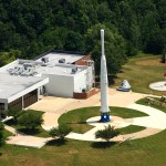 aerial view of a low-slung office building with a circular concrete sidewalk behind it, featuring a tall Delta A rocket in its center, with green grass and trees surrounding