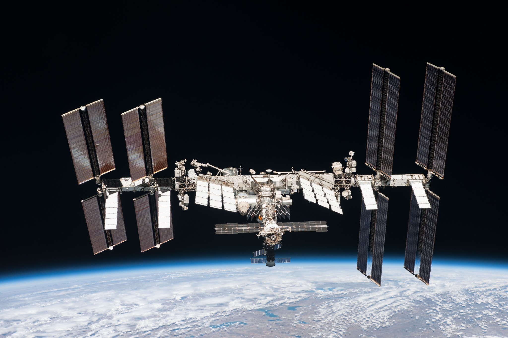 A photograph from space of the International Space Station orbiting Earth. The station is a graceful horizontal cylinder with four rectangular solar panels oriented vertically, two on either side of center. In the center are horizontal square panels angled slightly to the right, and science instruments and modules extend out from the center cylinder.