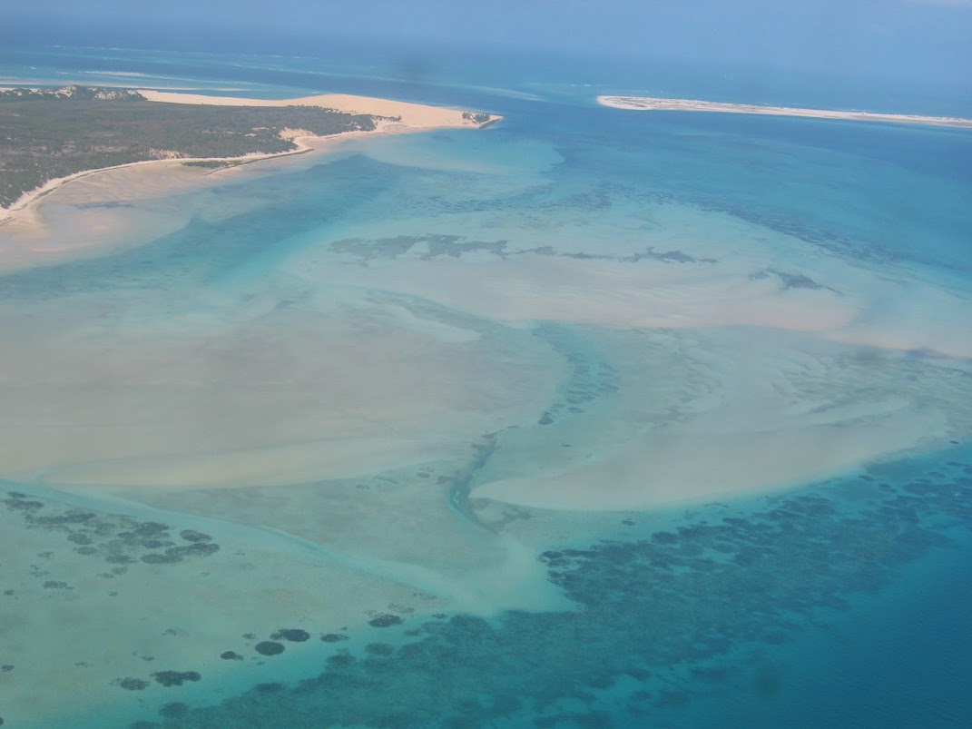 Aerial image off the coast of Mozambique. Bright blue ocean water covers a very shallow tan, sandy bottom, visible through the water. Dark patches of sea grass dot the edges of the sand bar.