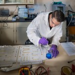 In a NASA lab, a male researcher in a white laboratory coat with latex gloves, dips a brush into a beaker filled with adhesive.
