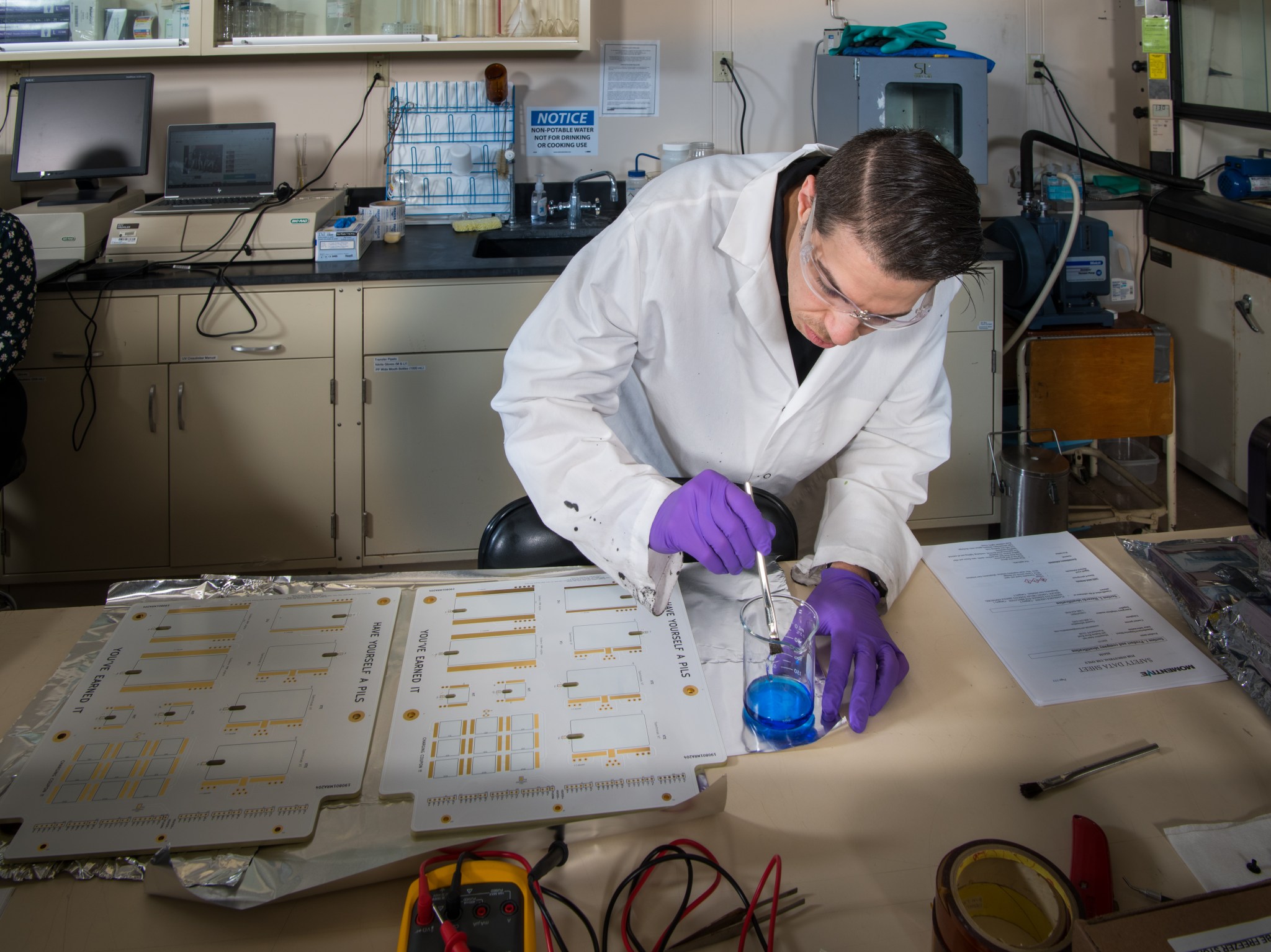 In a NASA lab, a male researcher in a white laboratory coat with latex gloves, dips a brush into a beaker filled with adhesive.