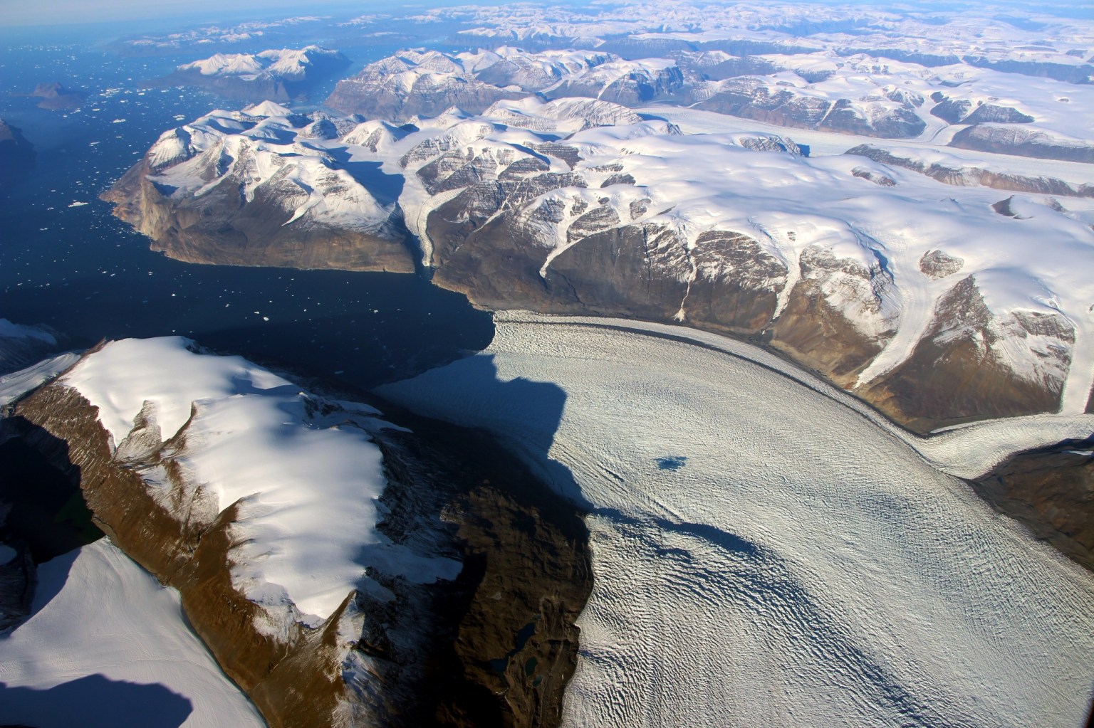 An aerial photo of a glacier in Greenland, looking like a wide, frozen gray-white river between two snow-mounded rock formations. The glacier has many small grooves and striations. The rocks on either side are tall and rounded, with smooth snow covering them like icing on buns. The rocky landscape extends out to the blue horizon, with deep blue water carrying small white ice floes in the left side of the image.