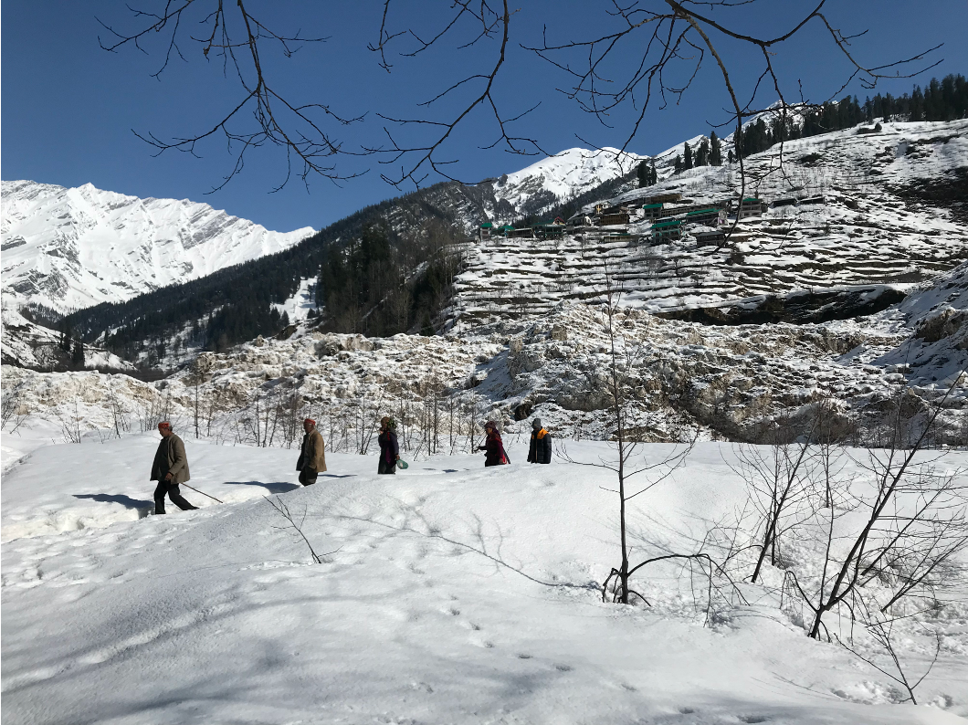 A group of people wearing winter gear walks through deep snow. Behind them, a rocky hillside has a lodge with green roofs built into the side. It is a bright, sunny day and the sky is deep blue.