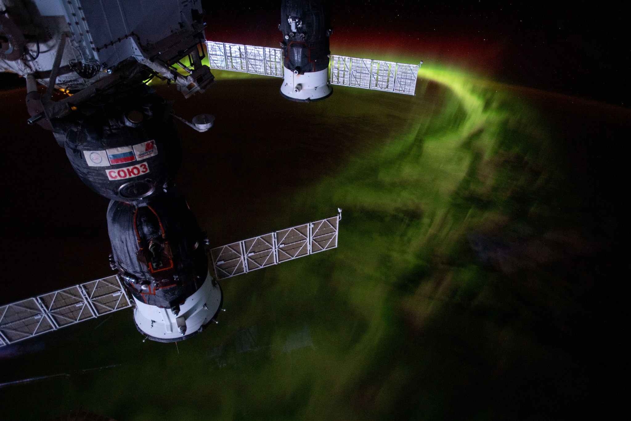 image of Russian space craft docked to ISS, with earth in the background, aurora australis (mostly green) visible