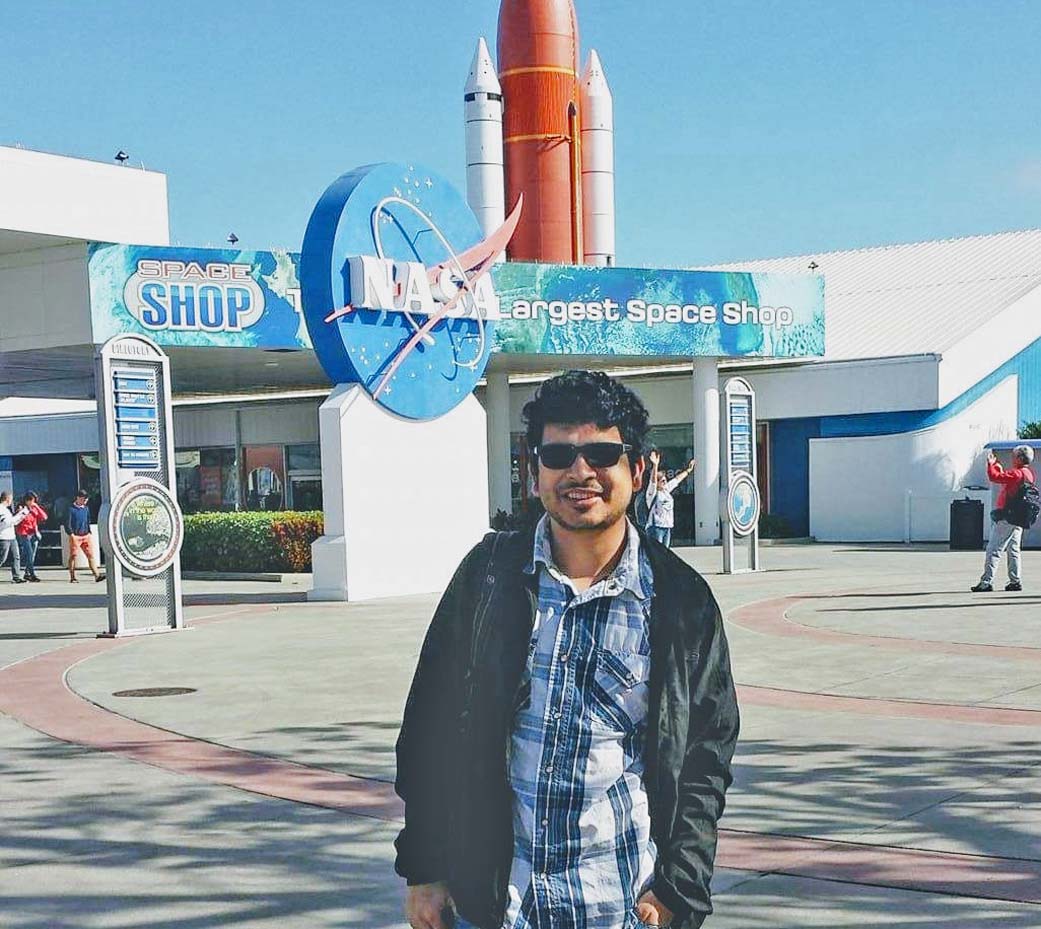 A man with black curly hair and tan skin wearing a blue plaid shirt, black jacket, black glasses, and a backpack, stands in front of the "Space Shop" with the NASA meatball and a large model rocket in the background at the Kennedy Space Center. 