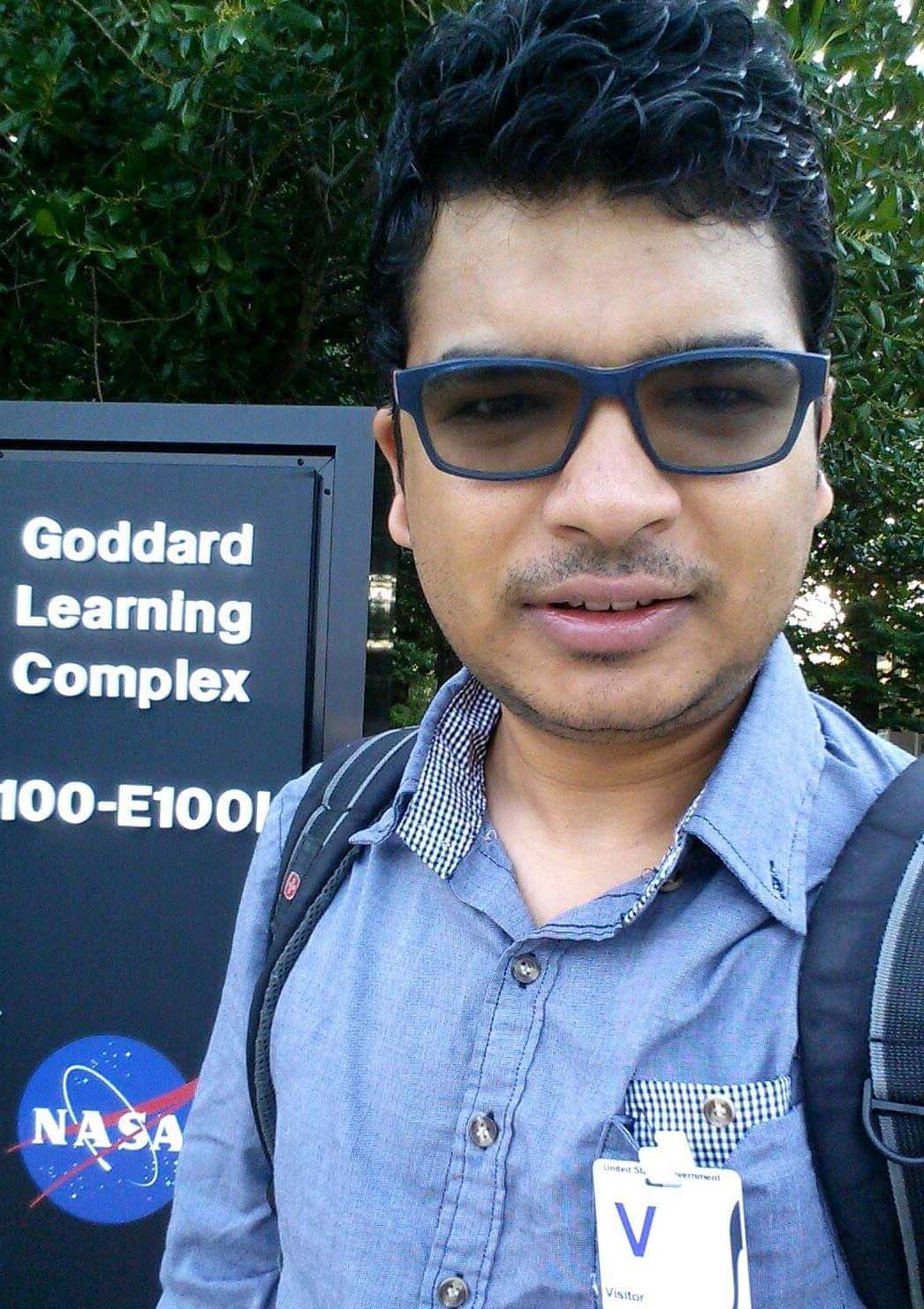 Man with black hair and tan skin wears black glasses, a blue collared shirt, and backpack.  He stands in front of a green tree and a sign that says "Goddard Learning Complex" with the NASA meatball