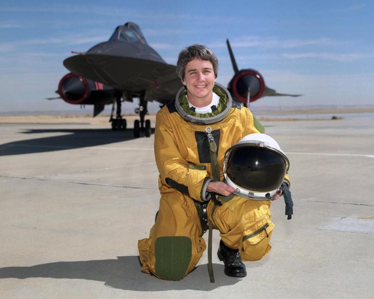 Woman in space suit holding helmet and posed in front of aircraft.