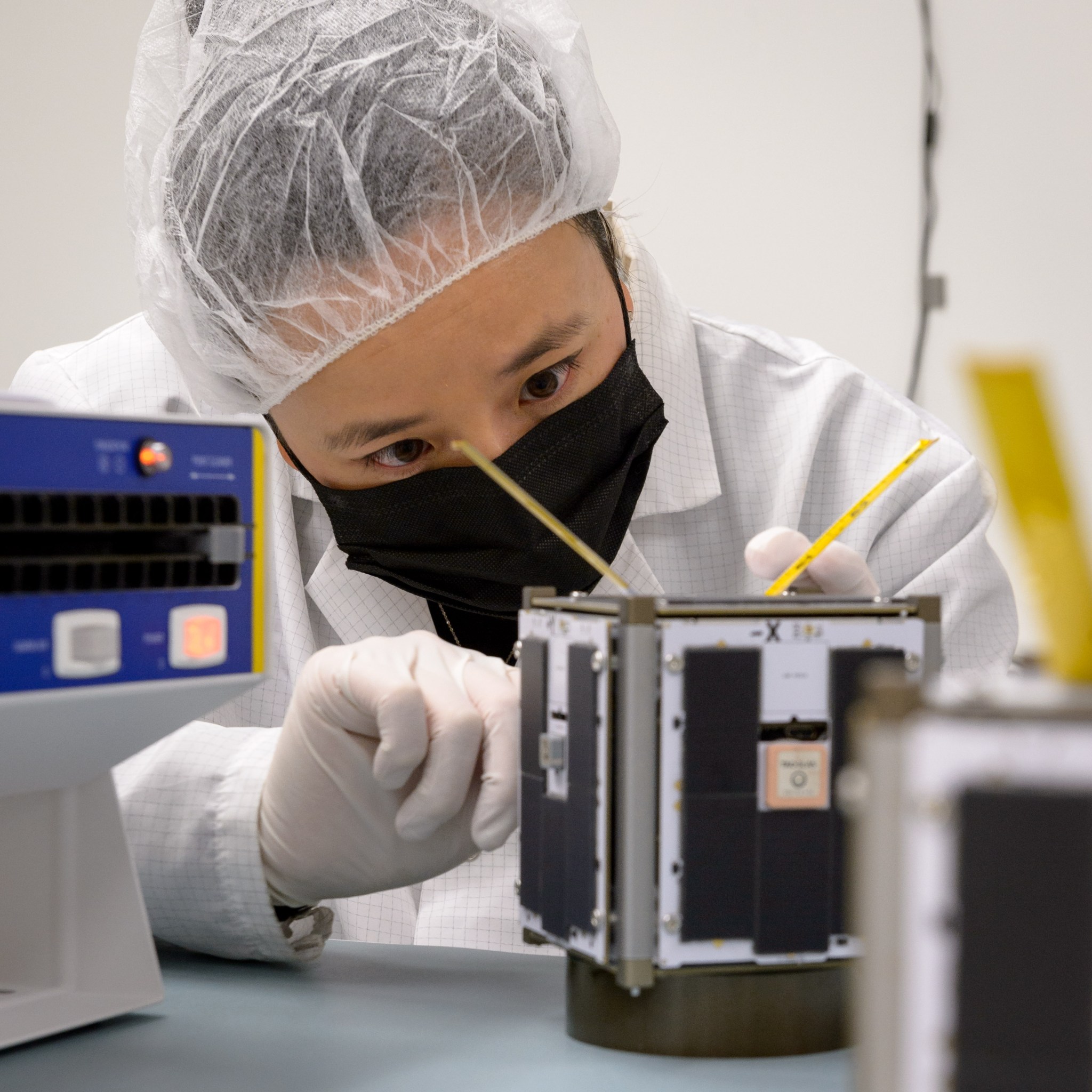 Person wearing a mask and hair net working on a small device.