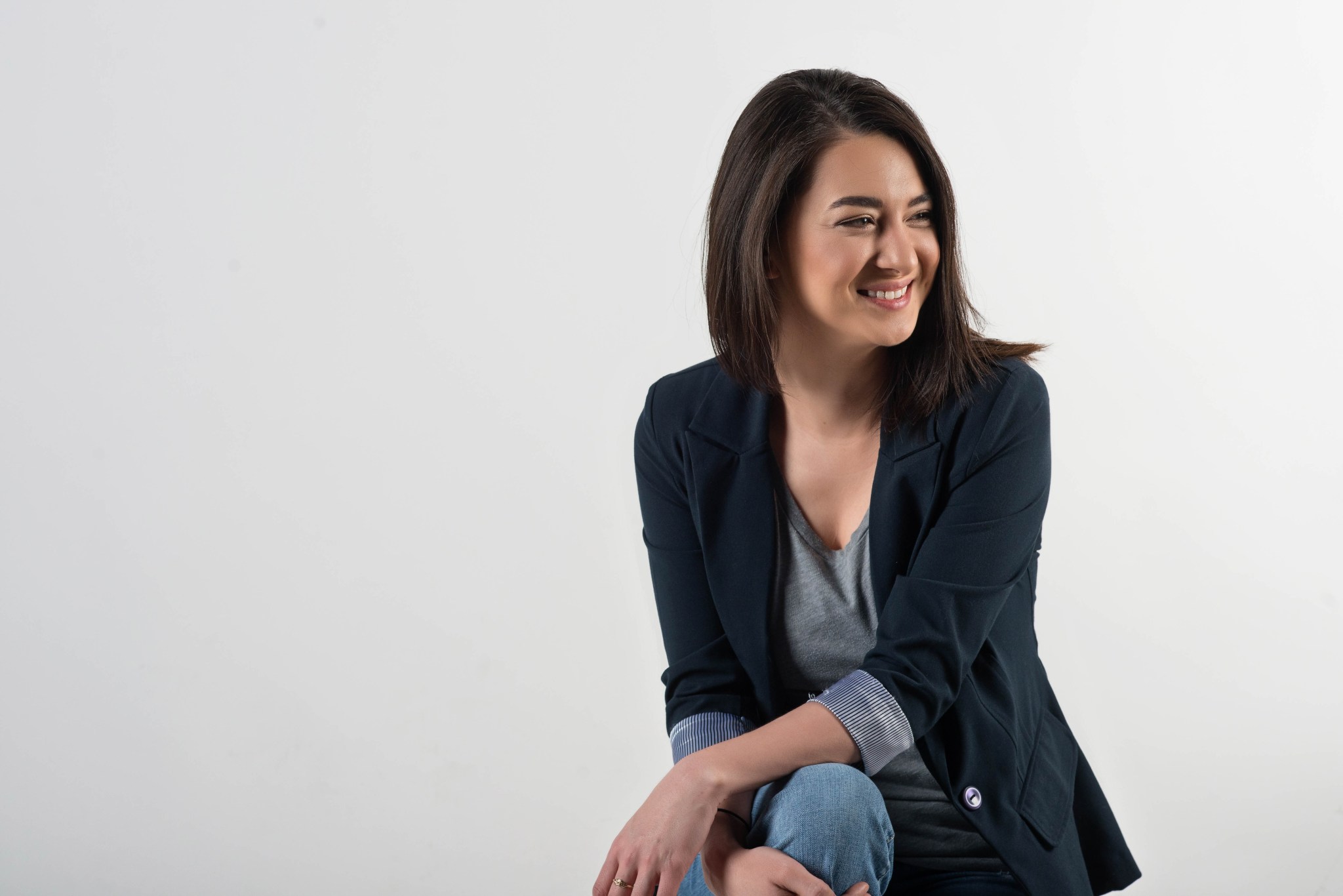 Woman with tan skin and dark brown hair looks off camera and smiles. She is sitting in front of a white background and wears jeans, a grey shirt, and a black blazer. 