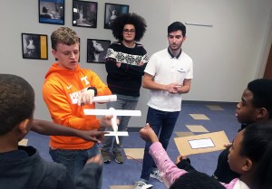 One student in a orange hoodie holding an airplane model with other students are pointing to it.