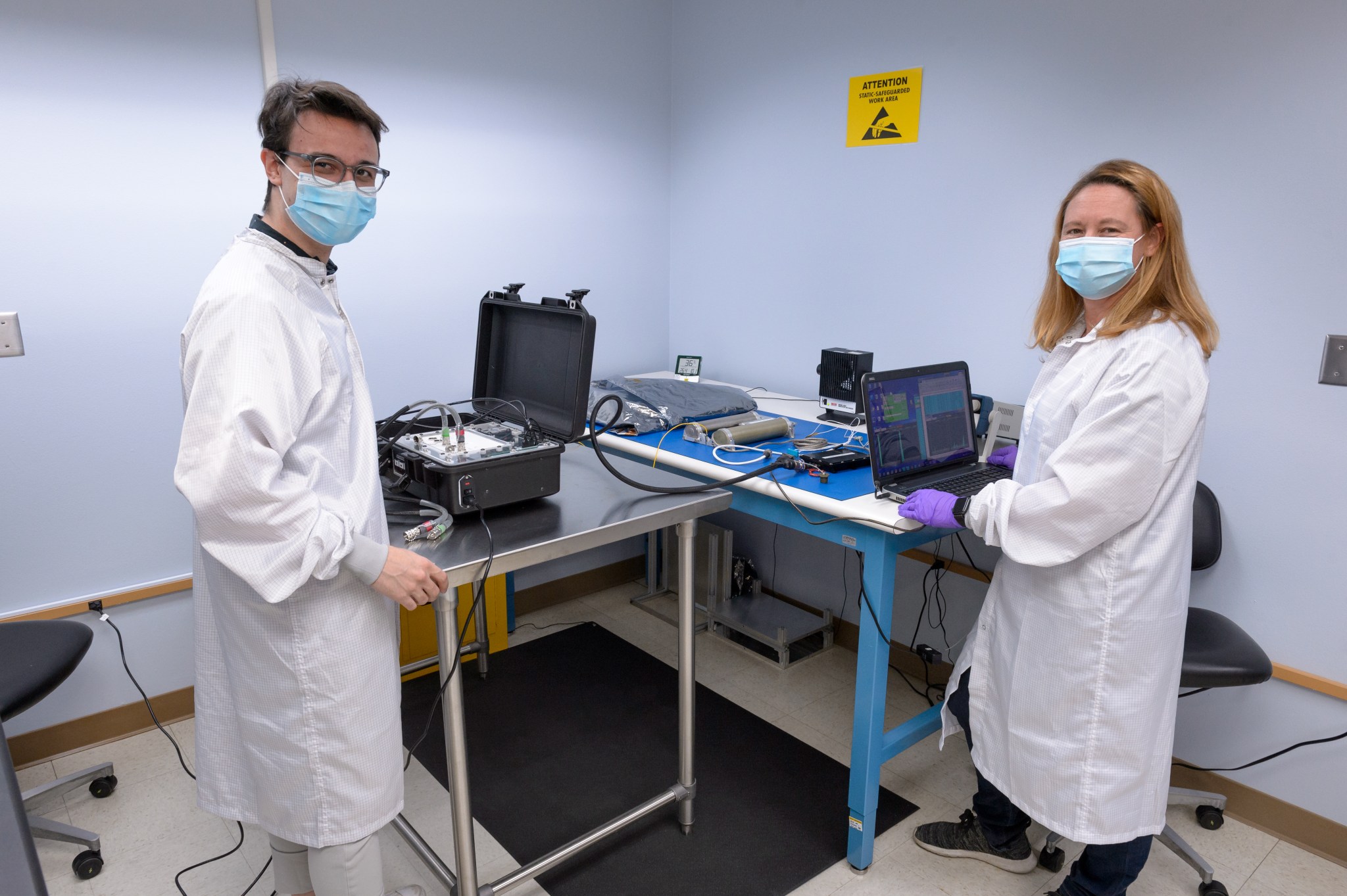 Two people in lab coats and masks standing over two connected tables, each with their own device connected by wiring.