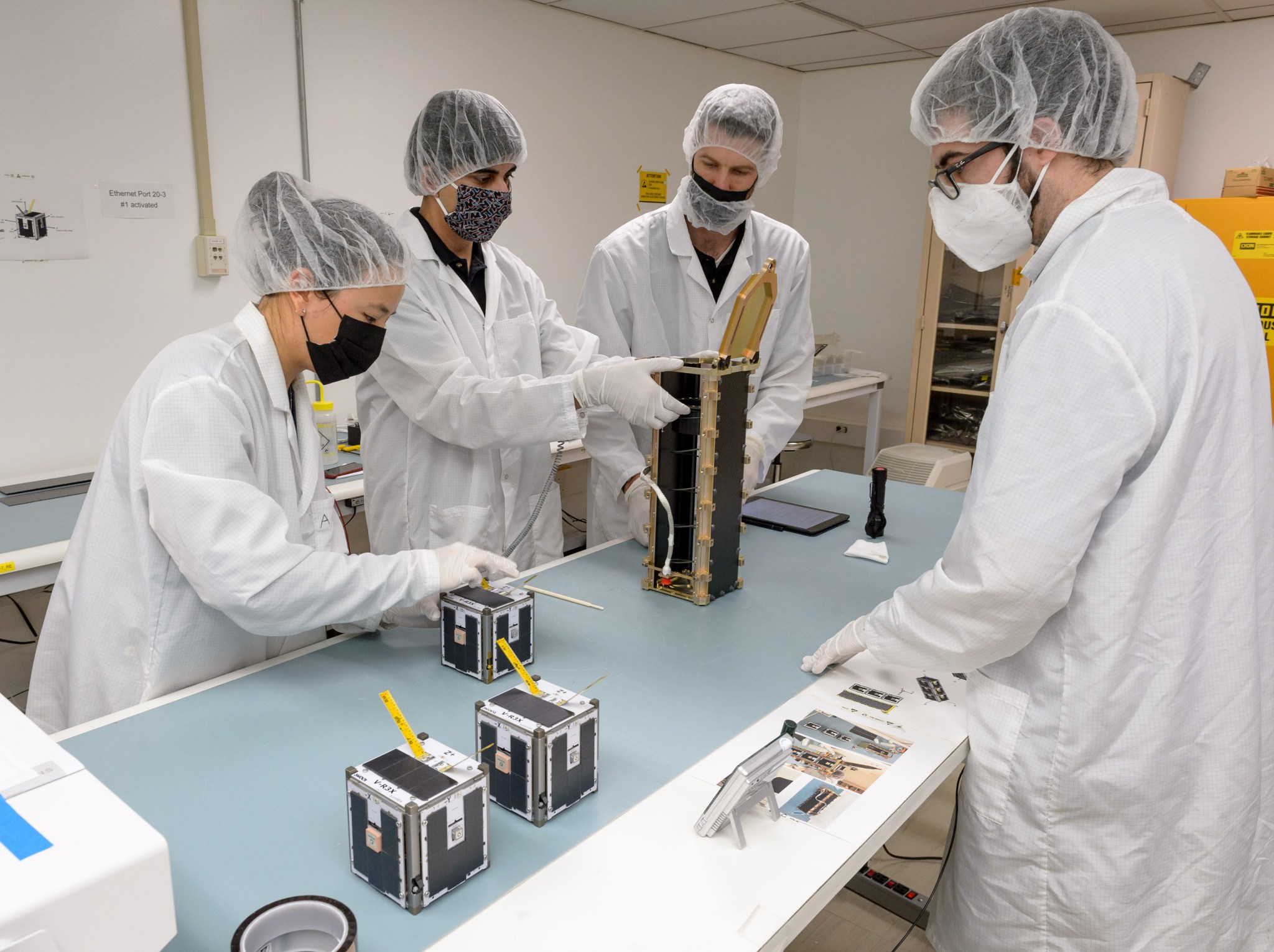 Four people standing around a table, with several small cube shaped devices and one longer, vertical container.