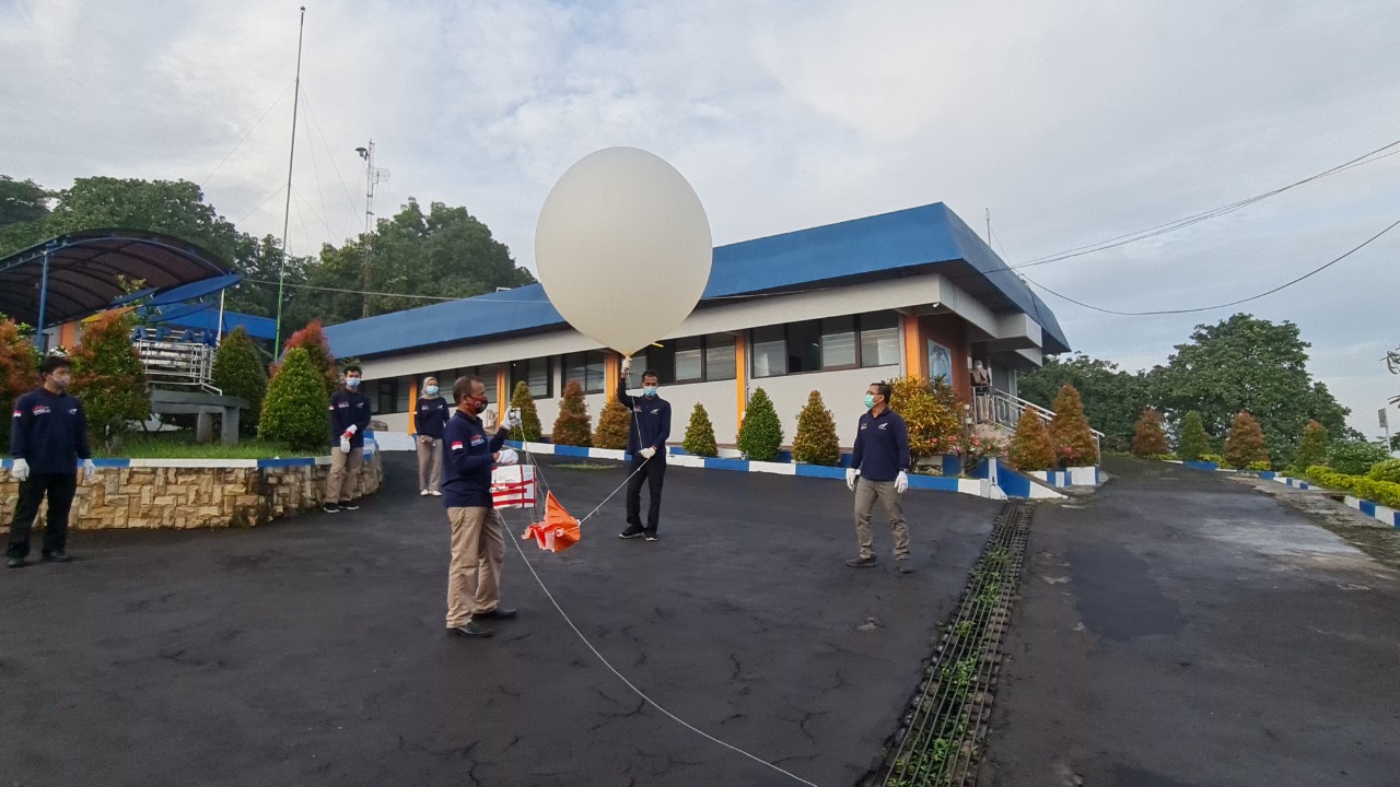 A group of scientists wearing blue jackets stands in a parking lot near a blue-roofed building, preparing to launch an ozonesonde, or large scientific balloon. Two people in the center hold the balloon, which is about as tall as a person and white, and has orange fabric and a long tether that extends off screen. Other scientists stand nearby and watch. Everyone wears masks and latex gloves.
