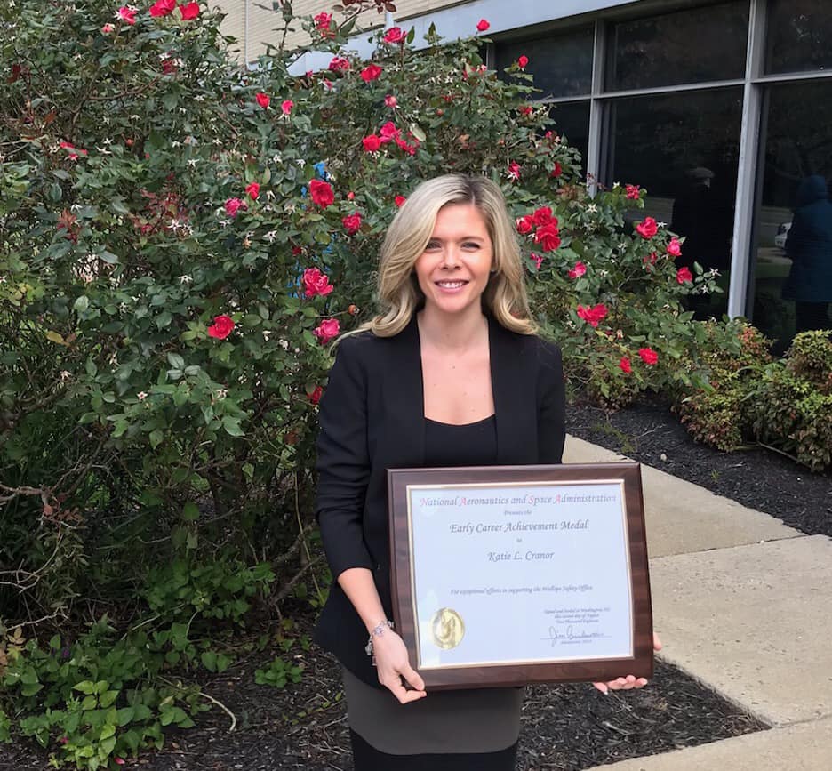 Woman with blonde hair wearing a black blazer stands in front of a rose bush holding a plaque that says "Early Career Achievement Award"