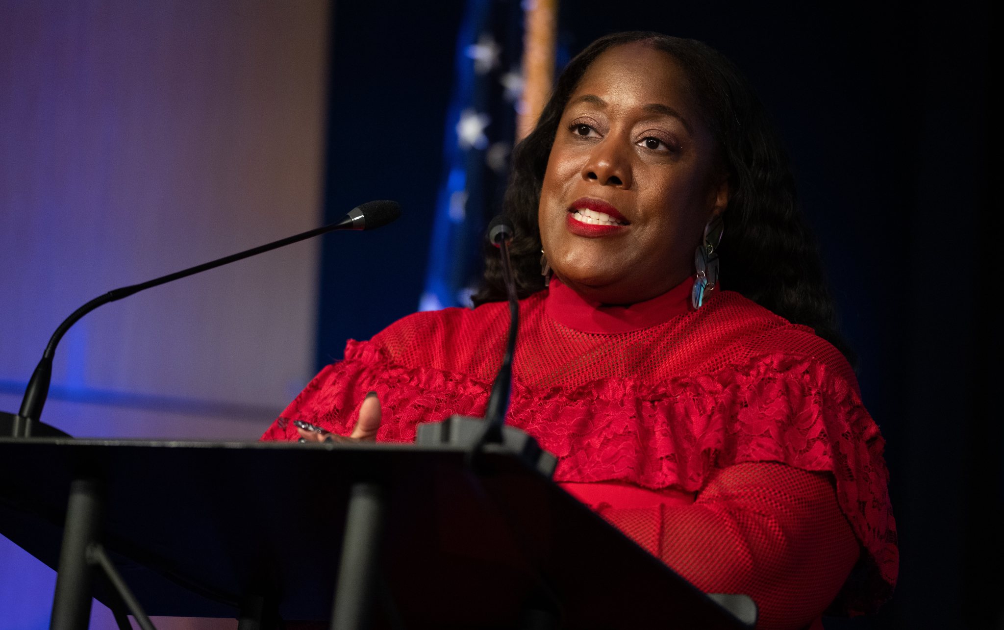 Michelle Jones stands at a lectern and speaks against an illuminated black and dark blue background. She wears a red blouse and iridescent earrings. An American flag is out of focus immediately behind her.
