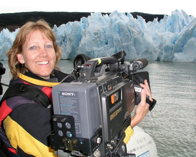Photo of a women holding a large video camera. A body of water and blue ice caps can be seen in the background.