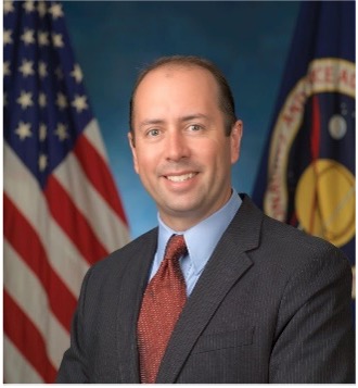 A smiling Eugene Schwanbeck in a charcoal suit with red tie with American flag and NASA flag in the background