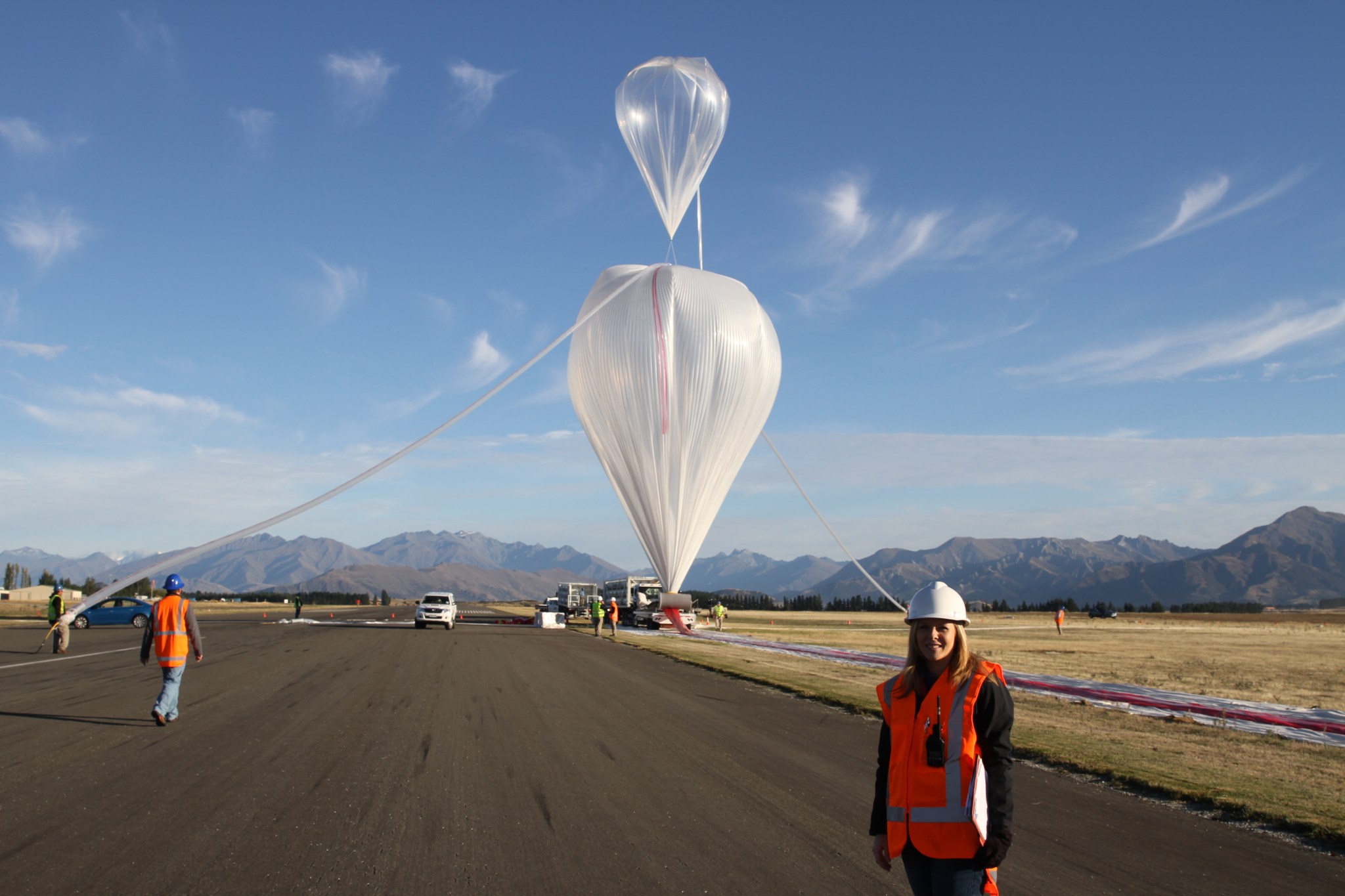 woman with blonde hair wears a hard hat and orange safety vest and is standing in front of two scientific balloons. The sky is blue with clouds and there are mountains in the background.